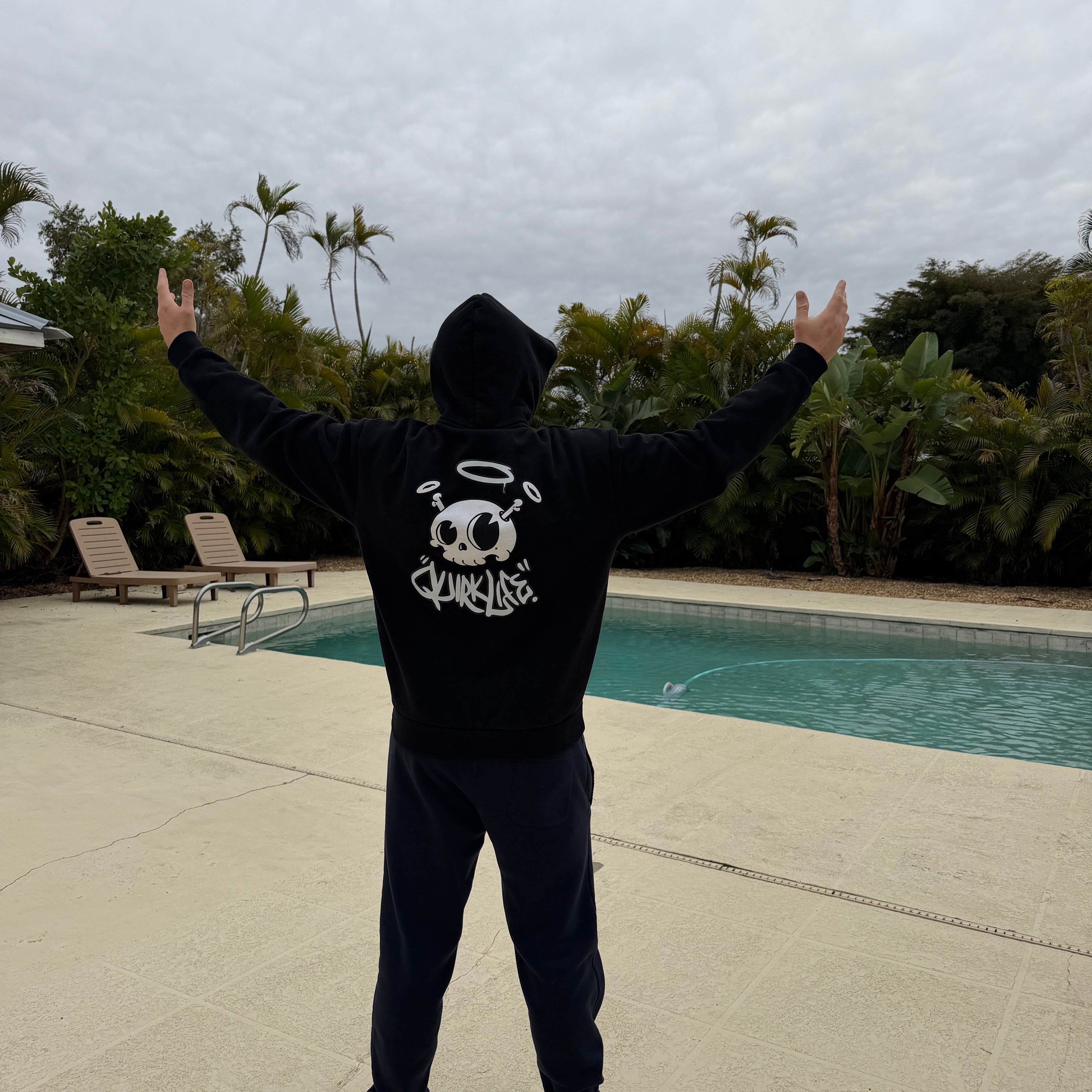 Person wearing a black hoodie with a graphic design, standing by a pool with palm trees in the background.